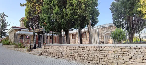 Taş duvar ve demir parmaklıkla çevrili tarihi yapı, büyük çam ağaçlarıyla süslenmiş bahçe ile çevrili.