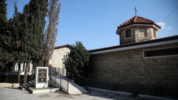 Mavi gökyüzü altında taş yapılı bir kilise, çatısında haç ve çevresinde düzenli doğa manzarası.