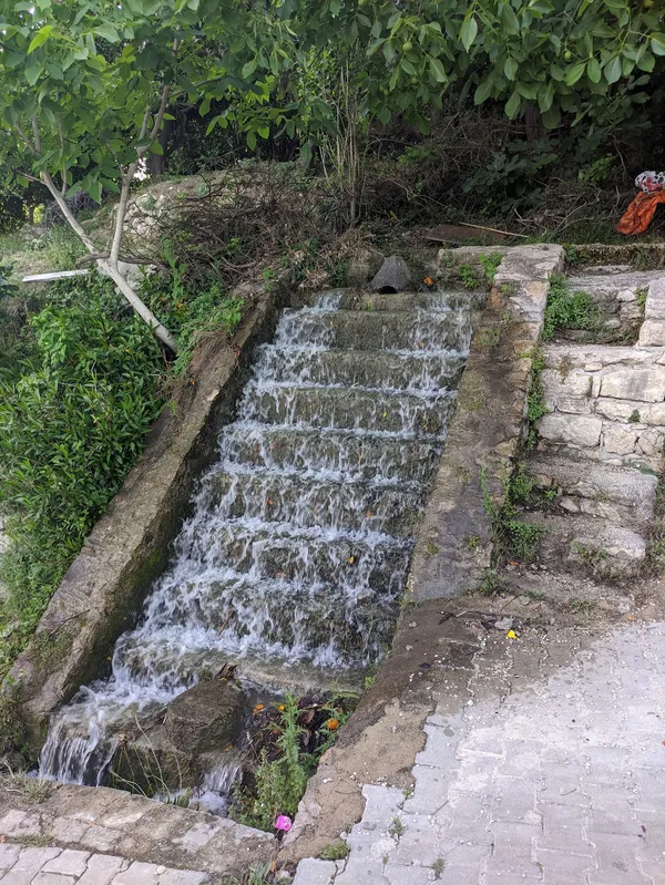 Yeşillikler içindeki şelale ve su kaynağı, taş yollarla çevrili huzurlu bir ortam sunuyor.