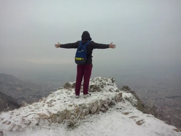 Karlı dağ zirvesinde, mavi sırt çantasıyla kollarını açan bir kişi, bulutlu manzaranın tadını çıkarıyor.