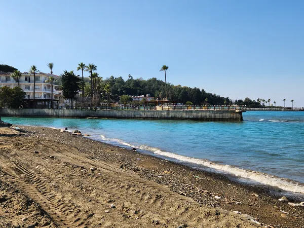 Arsuz plajında mavi su, kumsalda taşlar ve palmiyelerle çevrili oteller görünmektedir.