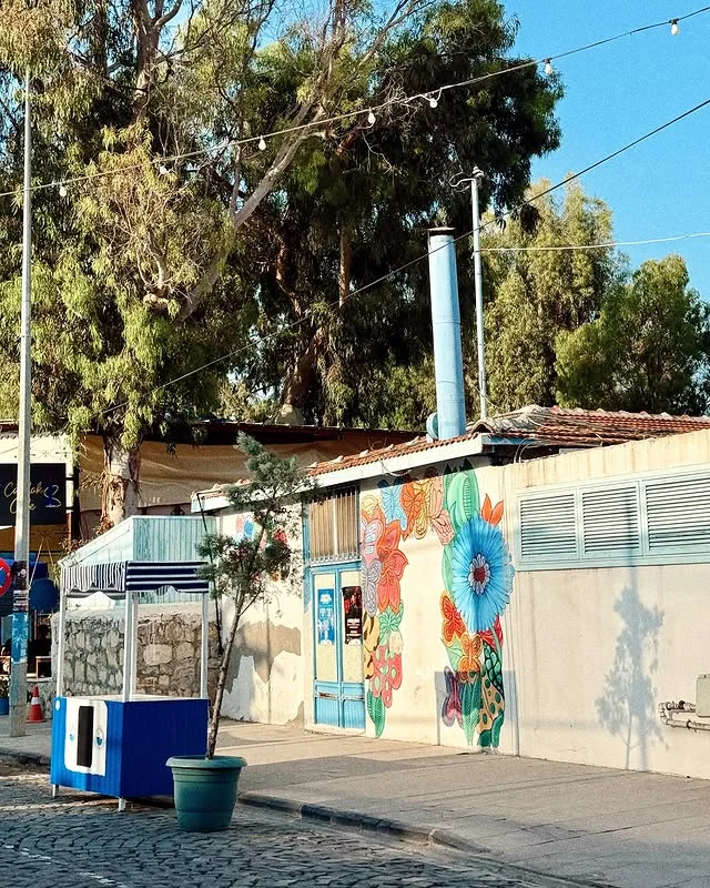 Arsuz'un Büyüleyici Renkleri - Mavi Kapı ve Çiçek Motifleri! Renkli çiçek motifleriyle süslenmiş bir duvar resmi, mavi kapılı yapı ve ağaçlarla çevrili.