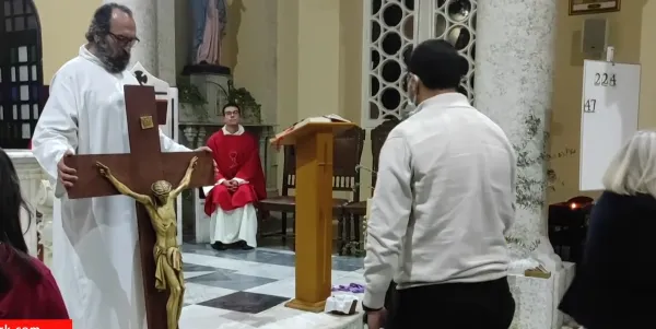 Beyaz elbiseli bir rahip, büyük haç tutarak dua ediyor, arka planda dini heykeller var.