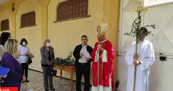 Kırmızı ve beyaz kıyafetli din adamı, sarı duvarlı kilise önünde grup insanla birlikte.