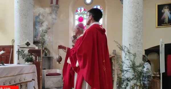 Kırmızı cübbeli erkeklerin katıldığı dini törende renkli vitray pencereler arka planda görünüyor.