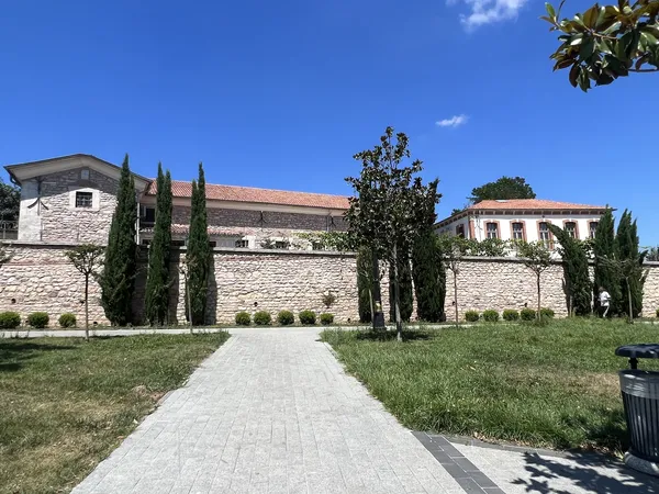 Yeşil alanda taş yolda selvi ağaçları ve kırmızı çatılı bir yapı görünmekte.