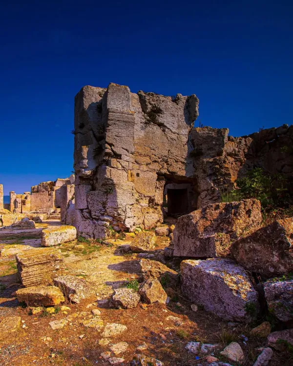 Yıkılmış antik yapının kalıntıları, mavi gökyüzü ve yeşil bitkilerle çevrili.