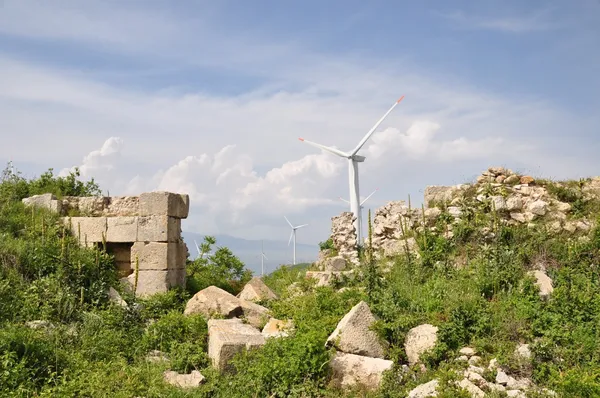 Yıkılmış taş yapılar, yeşil bitki örtüsü ve rüzgar türbinleri arasında güzel bir manzara.