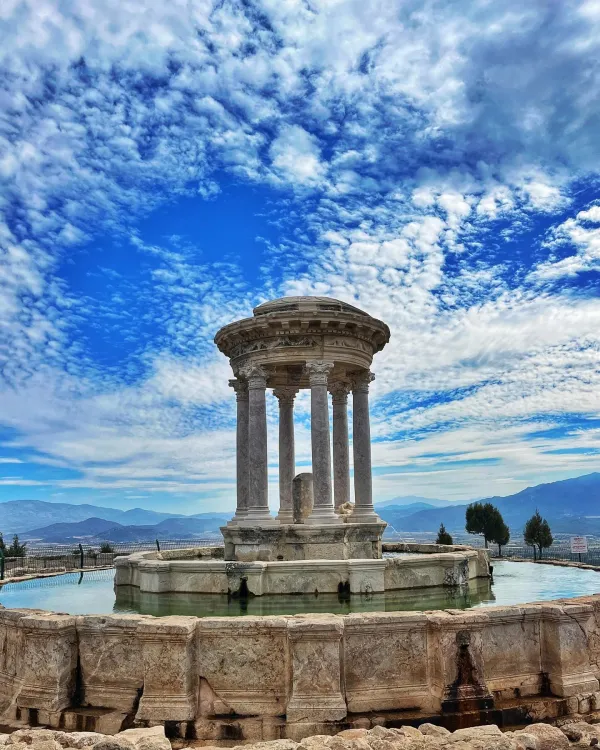 Yuvarlak üst yapılı tarihi bina, çevresinde su birikintisi, dağlar ve mavi gökyüzü ile doğal bir manzara sunuyor.