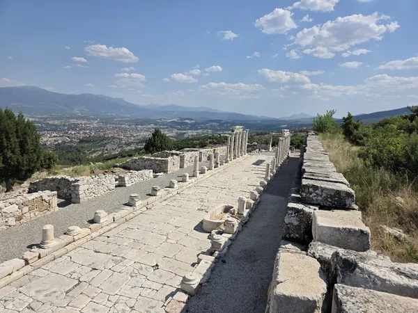 Geniş taş yol, yanlarında sütunlar ve arka planda taş duvarlar ile çevrili dağ manzarası.