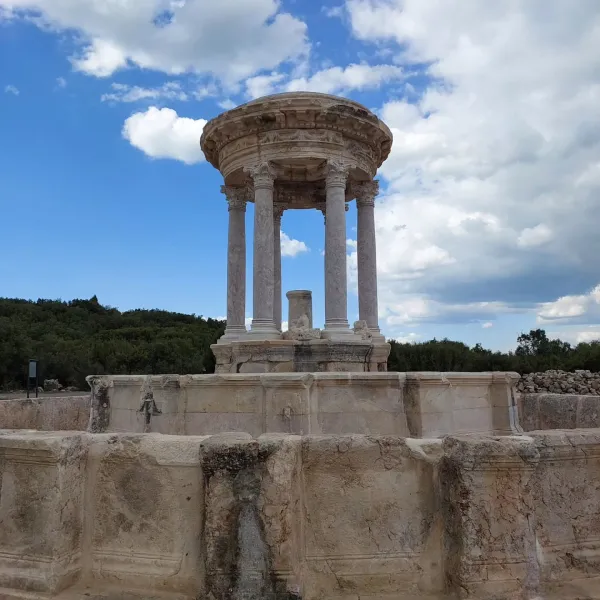 Sütunlarla desteklenen dairesel yapı, kubbesi ve yeşil ağaçlar arka planda.