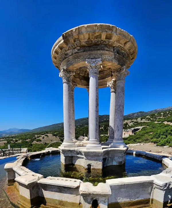 Dairesel yapıda sütunlar, kubbe ve su havuzu ile çevrili tarihi bir alan.