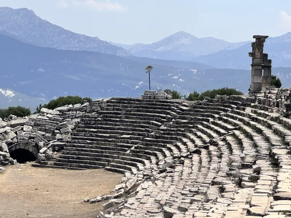 Geniş oturma alanı ve sahne kısmı ile antik tiyatro kalıntıları, arka planda dağlar ve mavi gökyüzü.