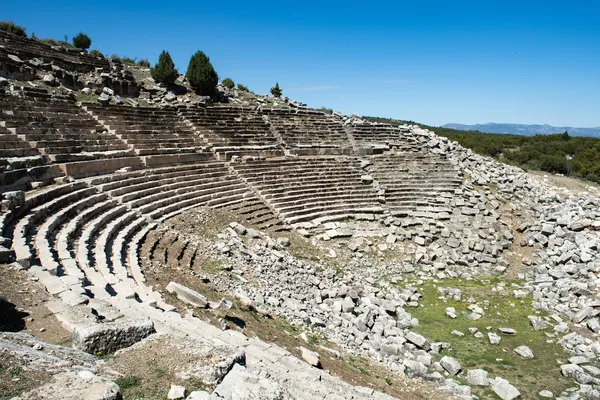 Kibyra antik kentindeki tiyatro kalıntıları, yeşil ağaçlar ve mavi gökyüzü ile tarihi bir atmosfer sunuyor.