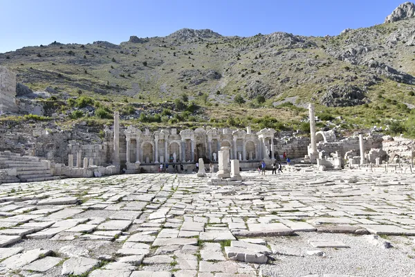 Sagalassos antik kentinde taş yapılar, sütunlar ve ziyaretçiler yer alıyor, arka planda yeşil dağlar var.