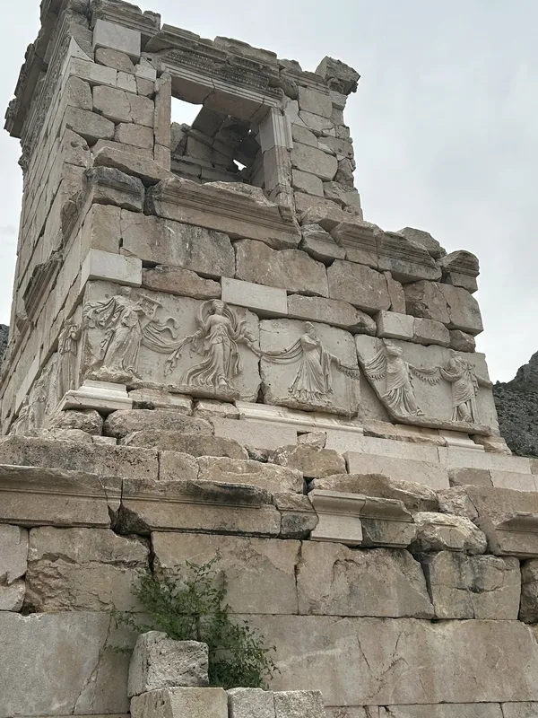 Taşlardan inşa edilmiş antik yapı kalıntıları, kabartmalar ve yeşilliklerle çevrili.