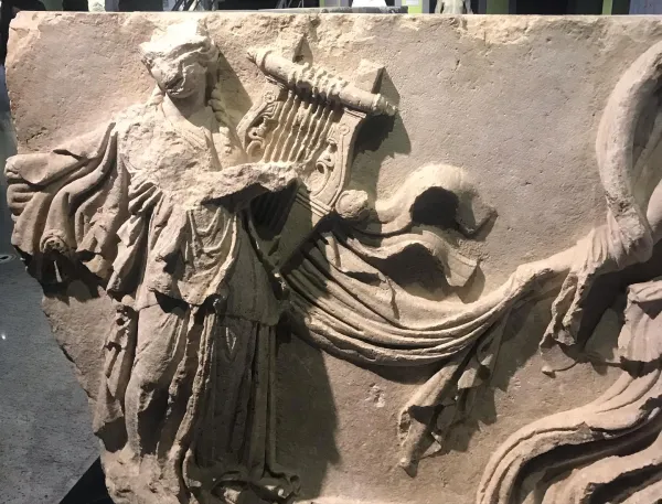 Antik bir yapıtta yer alan, elinde saz tutan ciddi yüz ifadesine sahip figür.