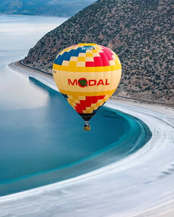 Rüzgarla süzülen renkli sıcak hava balonu, mavi-turkuaz Salda Gölü manzarasıyla birlikte