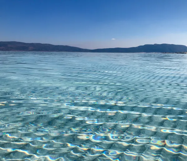 Berrak deniz yüzeyinde hafif dalgalar, arka planda dağ siluetleri ve açık mavi gökyüzü.