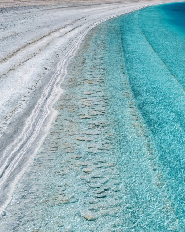 Berrak mavi su ve sade kumsal manzarası, dalgaların oluşturduğu kıyı çizgisiyle dikkat çekiyor.