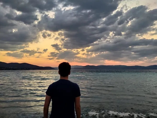 Deniz kenarında bir kişi, kararan gökyüzü ve dağlarla çevrili huzurlu bir manzaraya bakıyor.