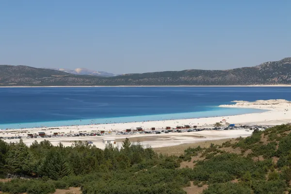 Geniş Salda Gölü manzarasında kumlu plaj ve berrak mavi su görünüyor, insanlar denizle eğleniyor.