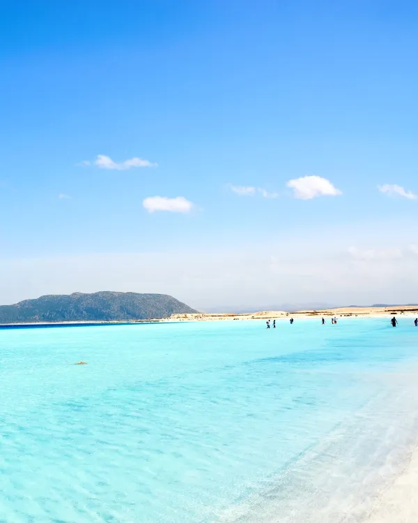 Masmavi suyu ve yeşil dağ siluetiyle Salda Gölü plajında insanlar yürürken.