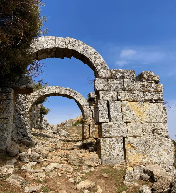 Yıkık dökük tarihi yapılar, mavi gökyüzü ve yeşil bitki örtüsü ile doğal bir manzara.
