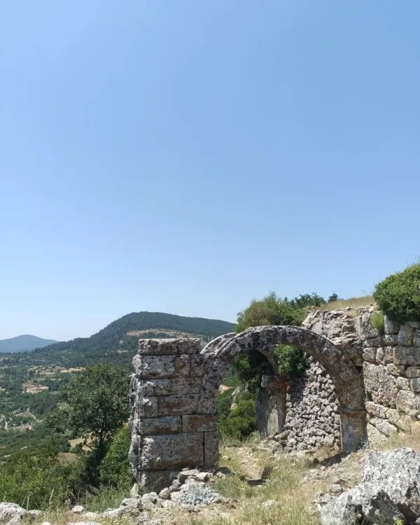 Antik taş yapının yuvarlak kemerli duvarı ve yeşil ağaçlarla dolu dağ manzarası