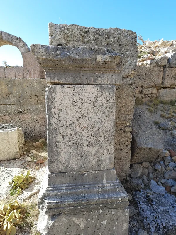 Aşınmış taş sütun, eski yapılar ve mavi gökyüzü ile çevrili.