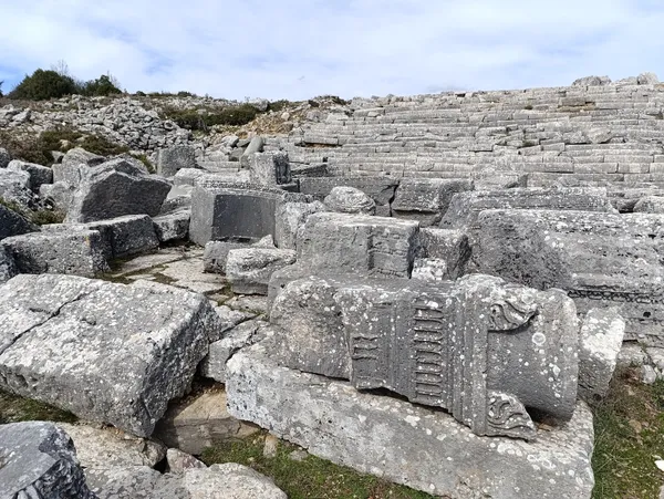 Yeşil zemin üzerinde dağılmış büyük taşlar ve eski tiyatro kalıntıları.