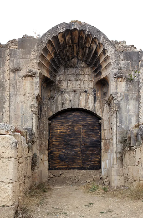 Büyük kemerli kapı, detaylı kabartmalar, taş duvarlar ve sarmaşıklar ile çevrili doğal bir zemin.