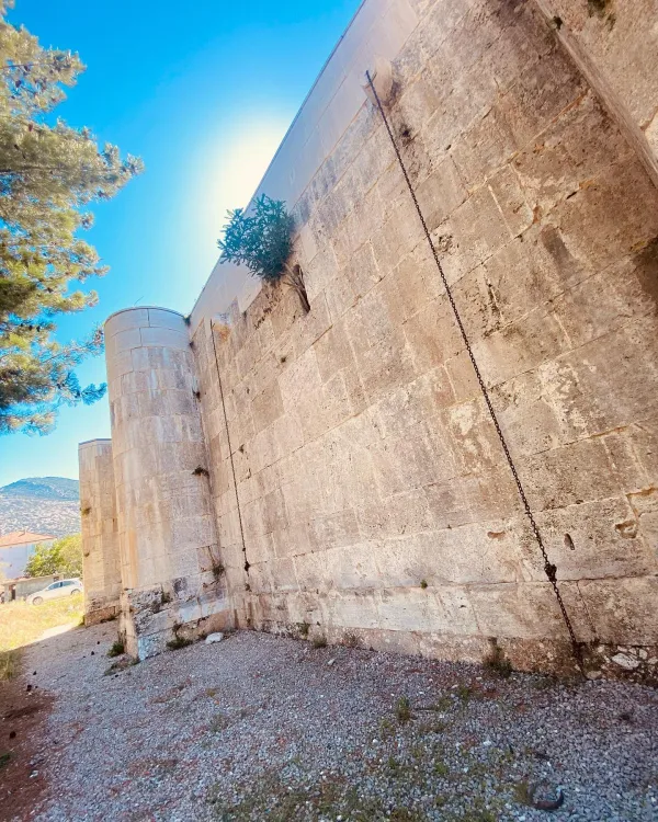 Büyük taş duvar, sarmaşıklarla kaplı, arka planda dağlar ve kırsal alan görünmekte.