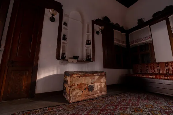 Koyu ahşap kapı, eski lambalar ve desenli halı ile taş oda konağı iç mekanı.