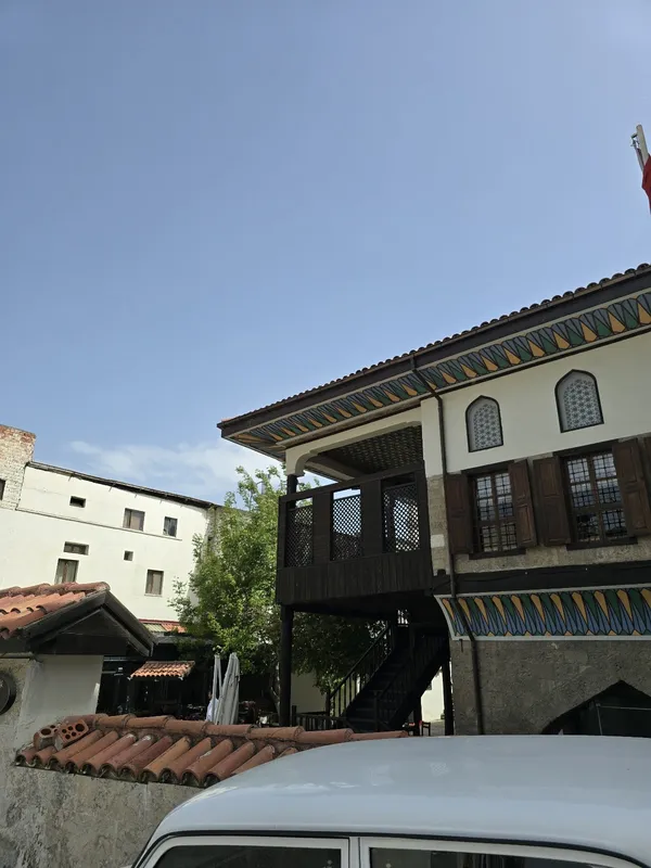 Geleneksel mimari tarzda taş oda konağı, ahşap pencereler ve yeşil ağaçlar ile çevrili.