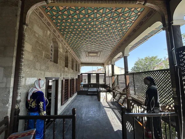 Geleneksel taş oda konağında balkon, ahşap detaylar ve yöresel kıyafetli mankenler.