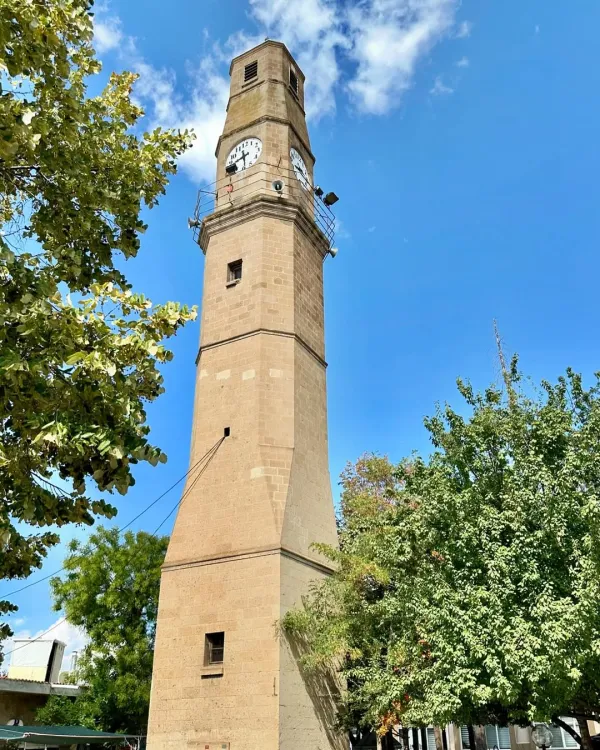 Taş yapılı saat kulesi, mavi gökyüzü ve ağaçlarla çevrili.