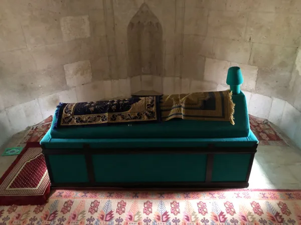 Taş duvarlarla çevrili alanda yeşil cenaze sandığı ve desenli örtüler