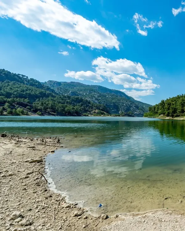 Sakin göl manzarasında yansıyan mavi ve yeşil tonlar, dağlar ve ağaçlar ile huzurlu bir atmosfer sunuyor.