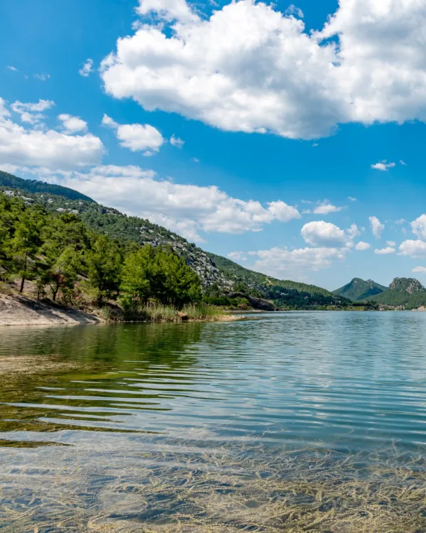 Sakin bir su kaynağı, yemyeşil dağlar ve açık mavi gökyüzü ile huzurlu bir manzara.
