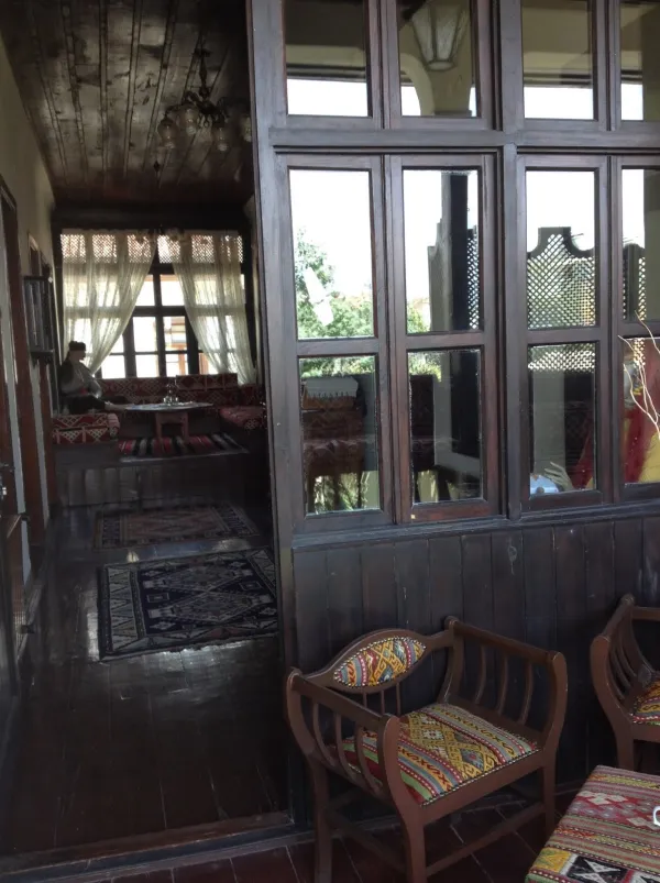 Geleneksel iç mekan, geniş cam pencereler, parlak ahşap zemin ve renkli halılarla süslenmiş oturma alanı.