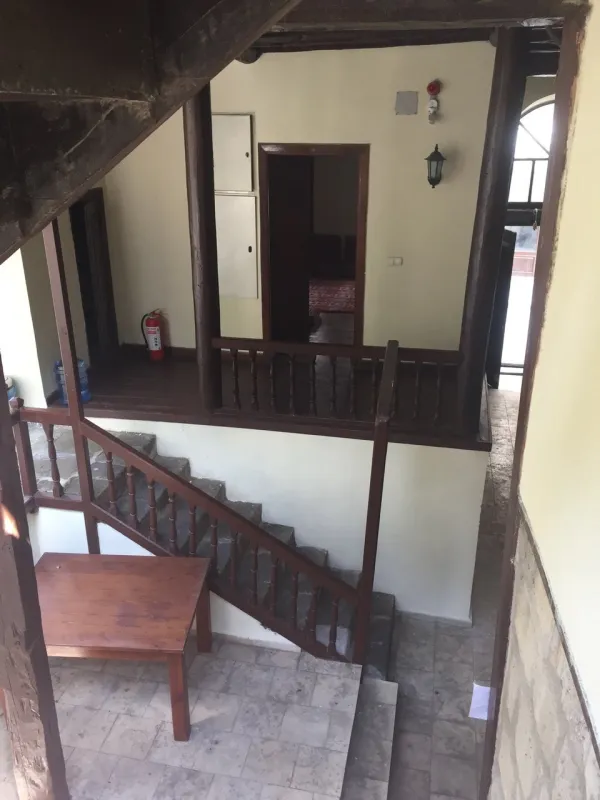 Ahşap merdiven ve açık alan içeren rustik bir mekan, beyaz duvarlar ve taş zemin ile aydınlık bir ortam sunuyor.