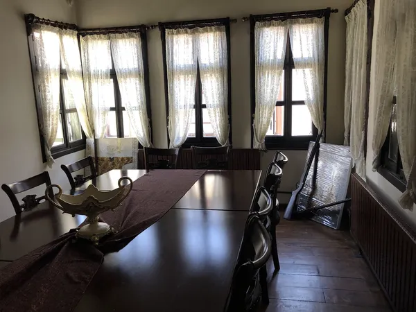 Uzun masa, dantel perdeler ve ahşap sandalyelerle aydınlık bir iç mekan.