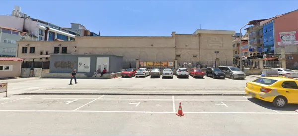 Geniş alanda büyük taş bina, park edilmiş araçlar ve mavi gökyüzü.