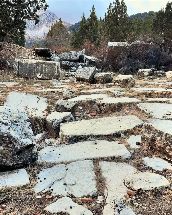 Büyük kare taşlardan oluşan zemin, kurumuş otlar ve dağlar arka planda.