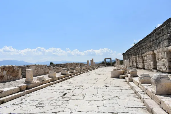 Geniş bir yol, antik sütun kalıntıları ve dağ manzarasıyla Balboura Antik Kenti.