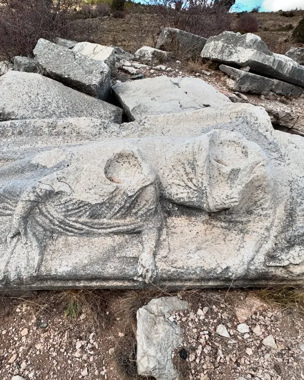 Büyük taşlar arasında, iki figürle süslenmiş heykel parçası doğal bitki örtüsüyle çevrili.