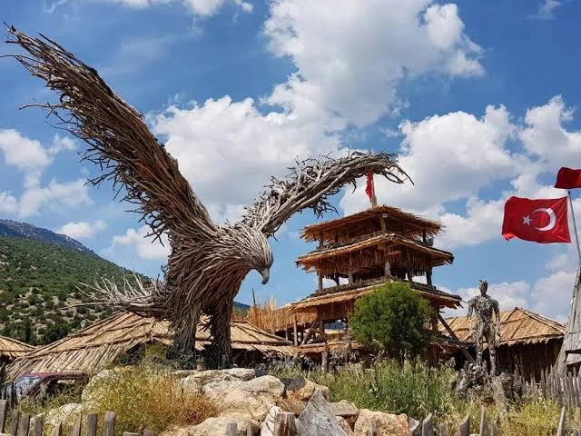 Lisinia Doğa Proje Alanı: Sanat ve Doğa Buluşması Açık kanatlarıyla detaylı kartal heykeli ve geleneksel yapı, yeşil bitkilerle çevrili.