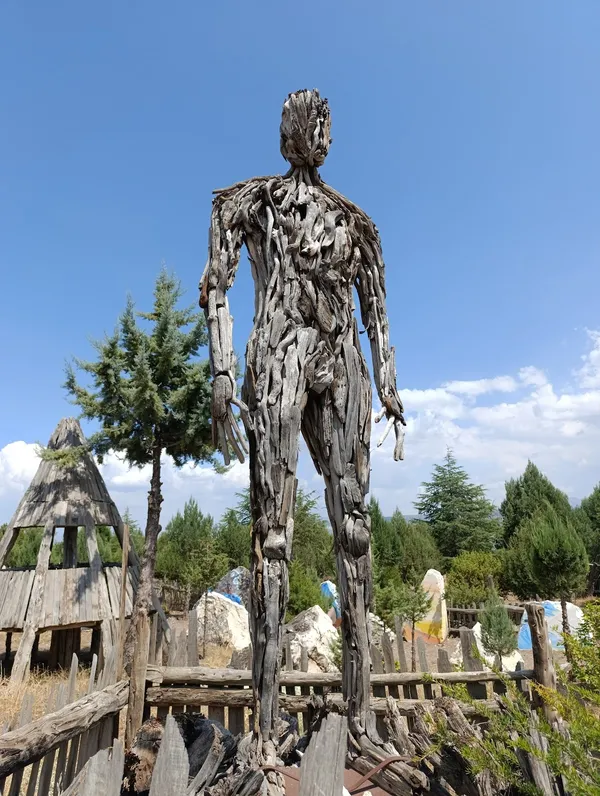 Büyük ahşap heykel, çam ağaçları ve eski yapı kalıntısı ile doğal ortamda yer alıyor.