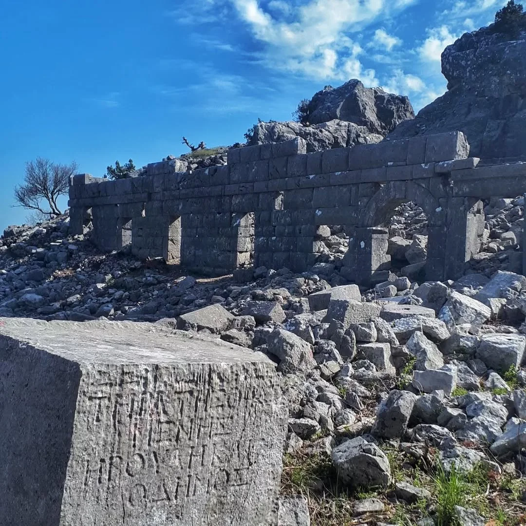 Milias Antik Kenti - Tarihin izleri ve doğal manzara! Milias Antik Kenti'nde taş bloklar ve kemerli duvar kalıntıları, arka planda doğal manzara ile birlikte.